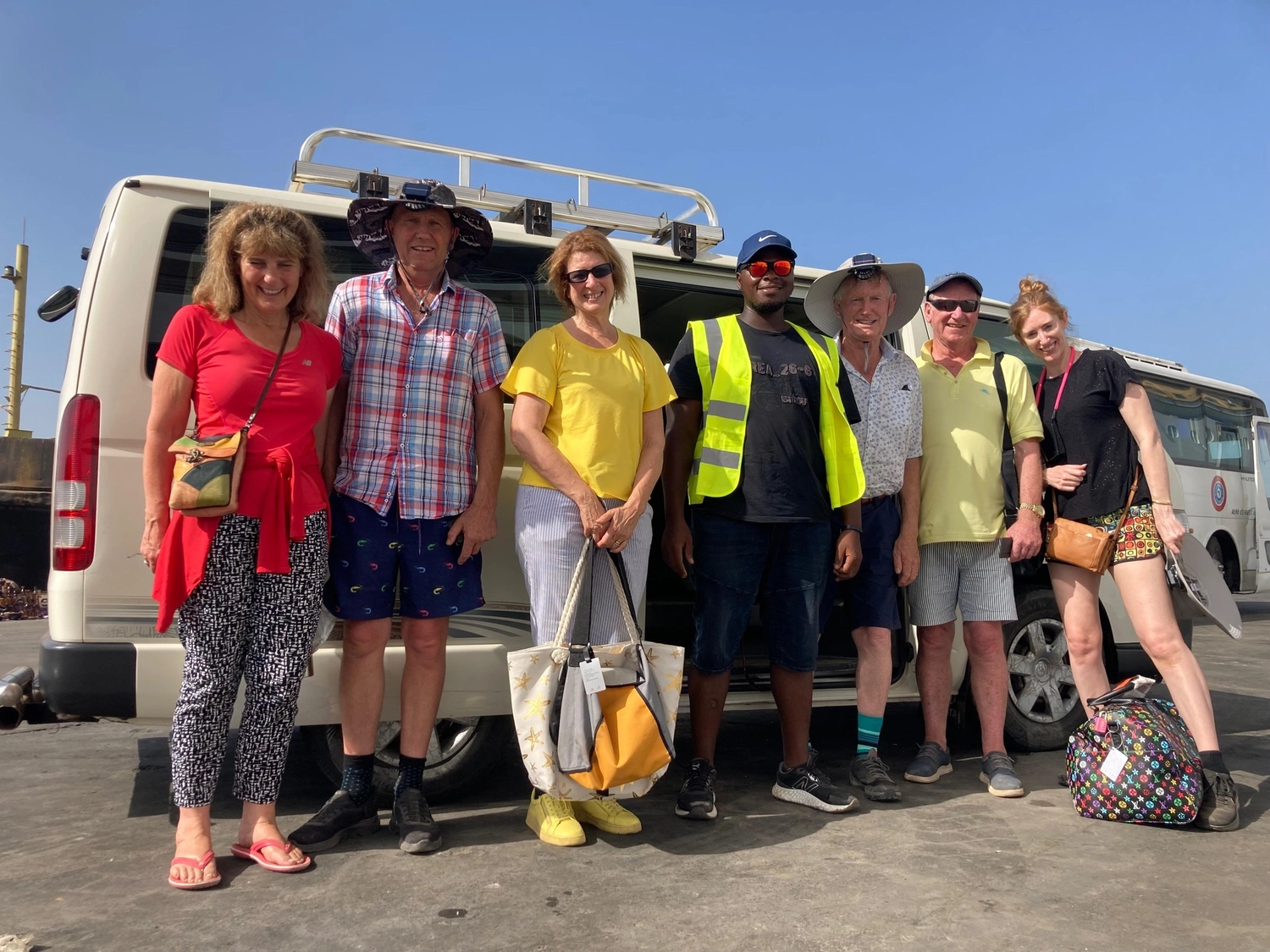 Gambia​ View Tours - Tourism services - A group of seven diverse tourists and a guide in a high-visibility vest standing together in front of a white tour van on a sunny day.