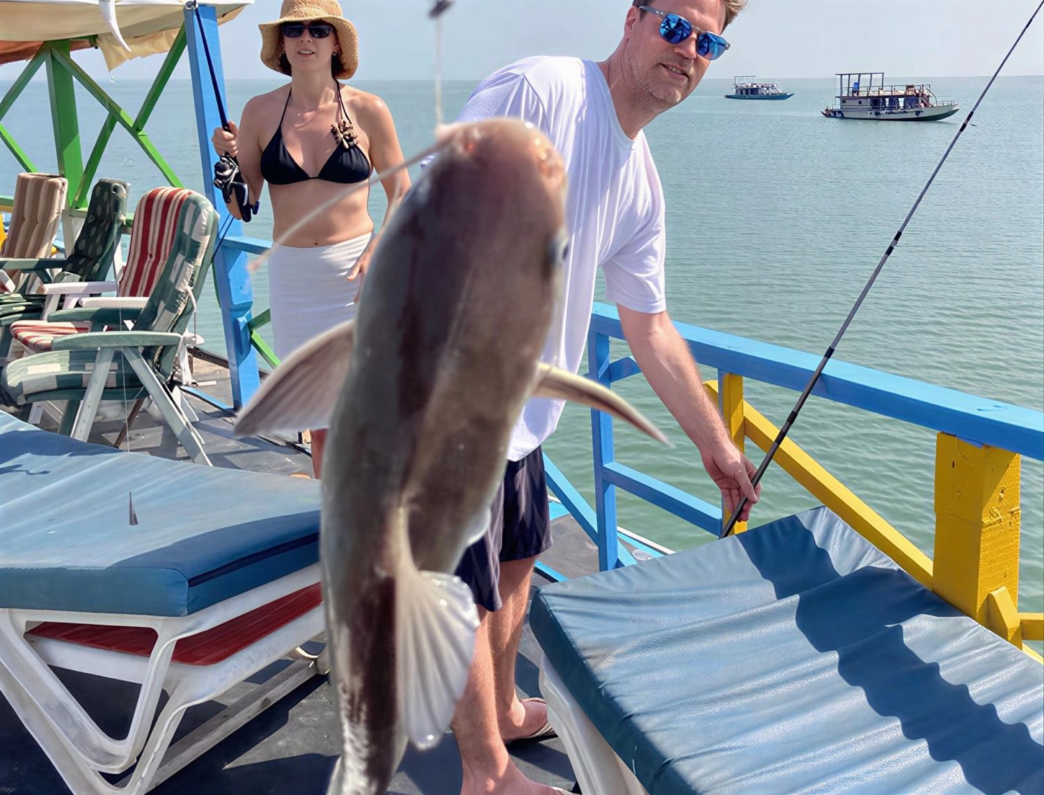 Gambia​ View Tours - Tourism services - A person holds up a fish they've just caught while standing on a brightly colored blue and yellow wooden pier by the water. Other people, lounge chairs, and a fishing rod are visible in the background.