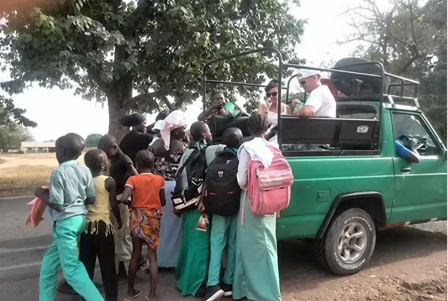 Gambia​ View Tours - Tourism services - A group of schoolchildren in green and orange uniforms gathered around a green safari-style truck as tourists look on from the back.